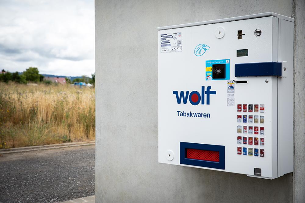 Ein Zigarettenautomat für den Außenbereich hängt an einer grauen Wand, im Hintergrund sieht man ein Feld.