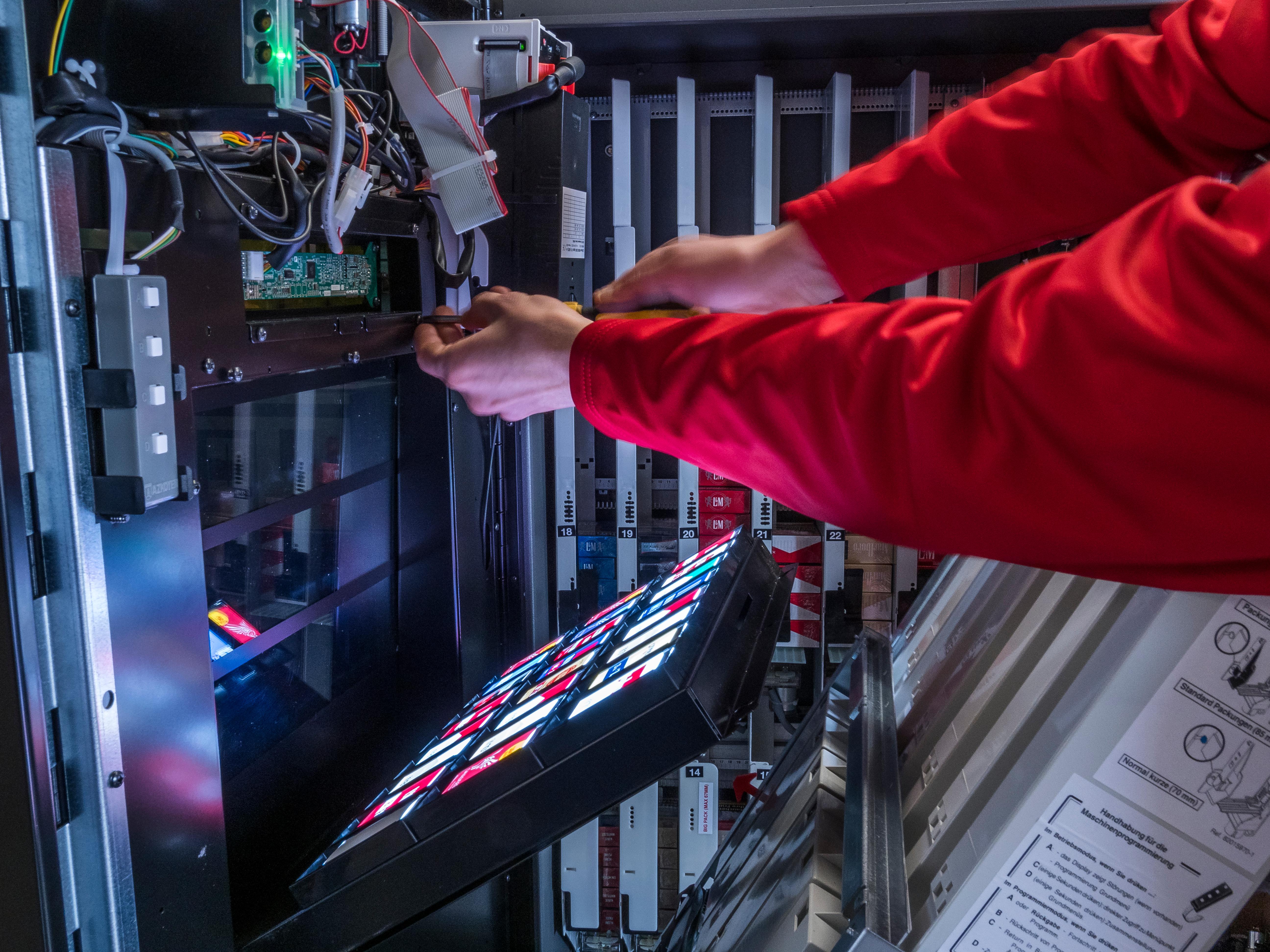 Automatentechnik Ein Automatentechniker schraubt an einem geöffneten, beleuchteten Zigarettenautomaten für den Innenbereich. Er trägt eine rote Jacke.