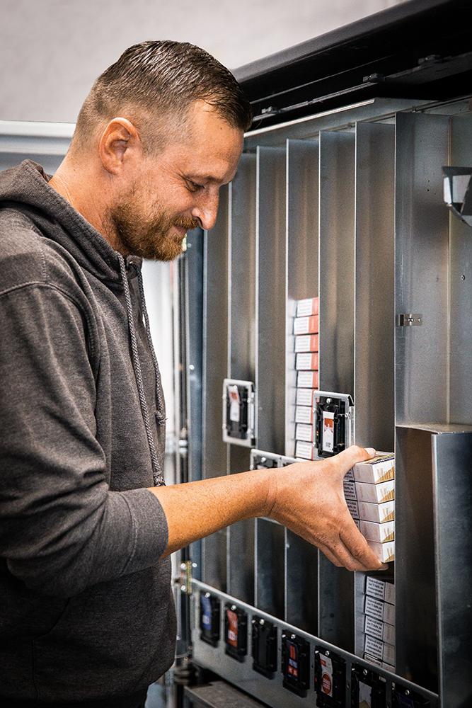 Ein junger Mann mit braunem Pullover füllt Zigarettenschachteln in einen Zigarettenautomaten.