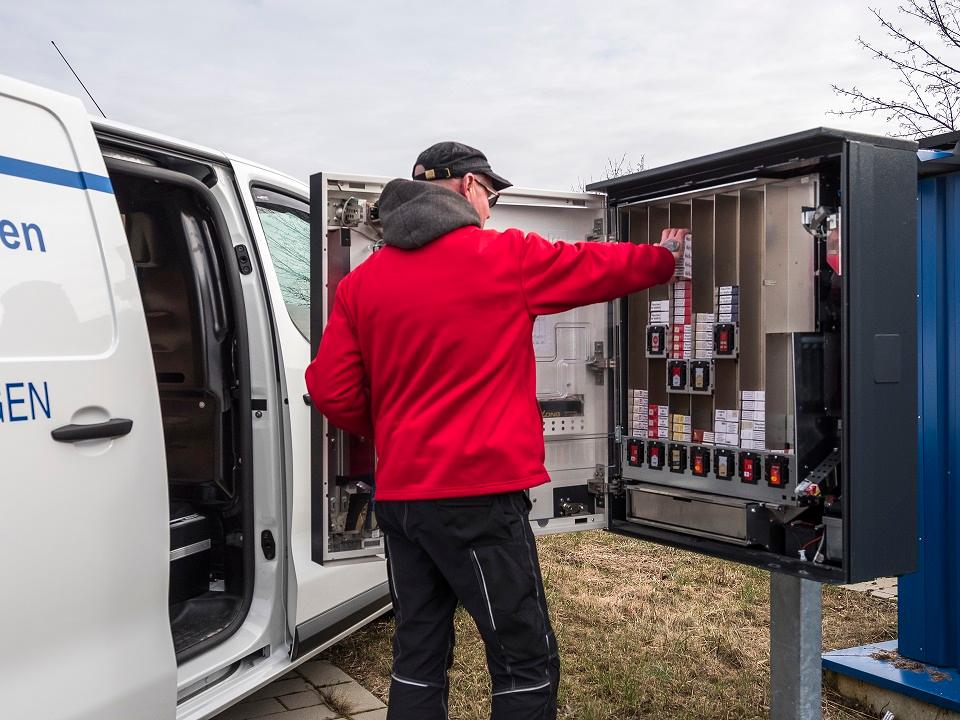 Ein Mann mit roter Jacke, der einen Zigarettenautomaten auffüllt und neben seinem weißen Lieferfahrzeug steht.