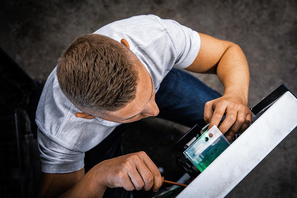 Techniker Ein junger Mann mit einem grauen Poloshirt, der an einer technischen Komponente eines Zigarettenautomaten schraubt, von oben.