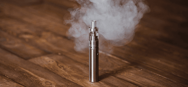 Ein silbernes Vape-Gerät liegt auf einer dunkelbraunen Holzoberfläche und stößt aufsteigenden weißen Rauch aus.