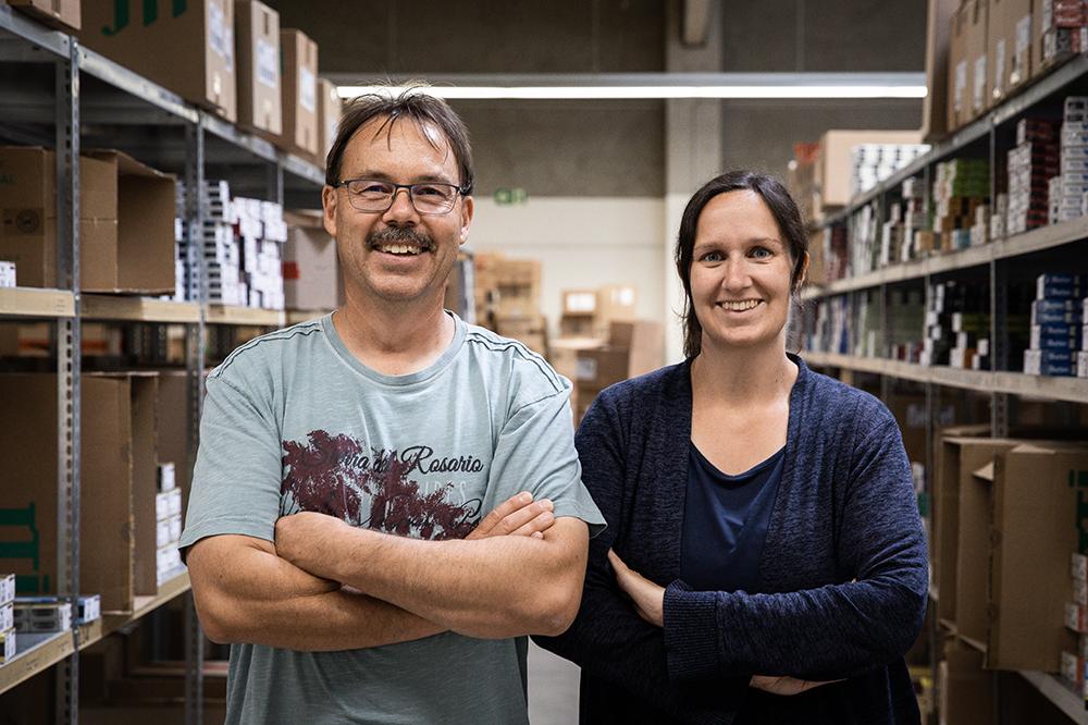 Links ein Mann mit blauem Shirt und rechts eine junge Frau mit blauem Pullover, die vor Lagerregalen stehen.