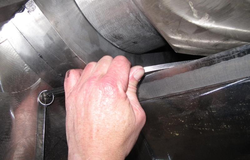 Eine Hand misst mit einer Fühlerlehre eine Metalloberfläche – den Verschleiß des Mischers –, umgeben von glänzenden Metallteilen.