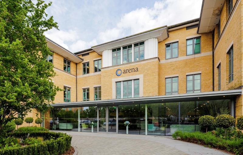 A modern, tan-brick office building with large windows and a sign reading "arena" above the entrance, surrounded by neatly landscaped greenery.
