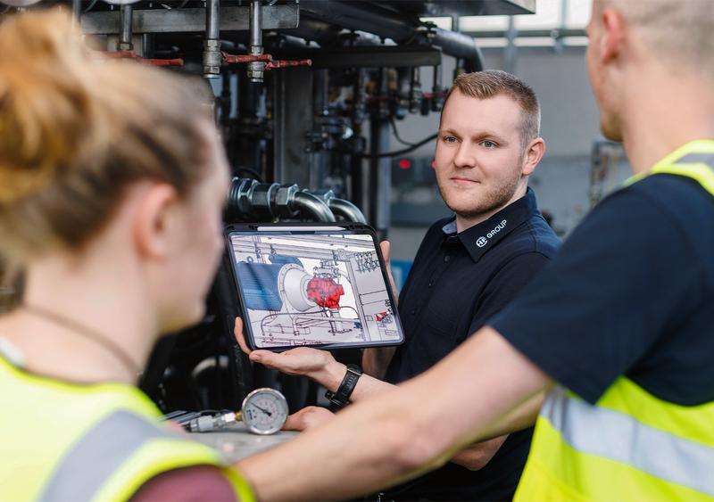 Technician shows two colleagues a digital plant model on a tablet, standing among pipes and valves in an industrial production environment.